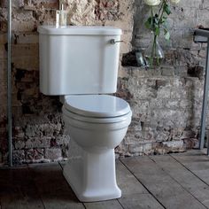 a white toilet sitting on top of a wooden floor next to a brick wall and potted plant