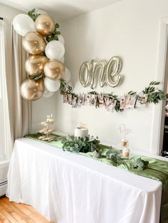 a white table topped with balloons and greenery next to a sign that says one