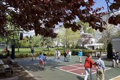 people playing tennis on an outdoor court in a park with lots of trees and benches