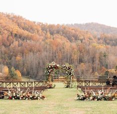 an outdoor ceremony set up with wooden benches and floral arrangements in front of fall colored trees