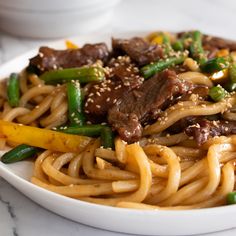 a white bowl filled with noodles and beef on top of a marble countertop next to chopsticks