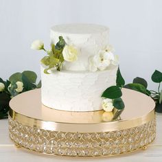 a wedding cake with white flowers and greenery on the top is sitting on a gold platter