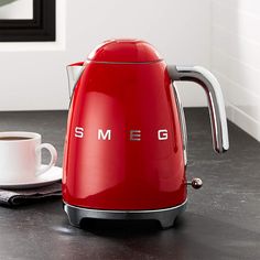 a red smeg kettle sitting on top of a counter next to a cup of coffee