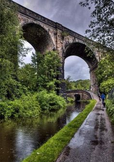 a man walking down a wet road next to a river under an old stone bridge