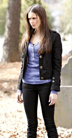 a young woman standing in the leaves wearing black pants and a blue shirt with a black jacket over her shoulders