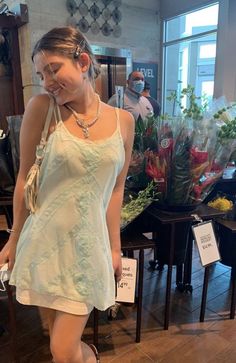 a woman is standing in front of flowers at a flower shop wearing sandals and a white dress