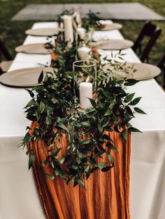 a long table with candles and greenery on it is set for an outdoor dinner