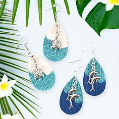 three pairs of earrings with dolphins on them sitting next to palm leaves and white flowers