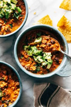 three bowls of chili with tortilla chips and avocado on the side