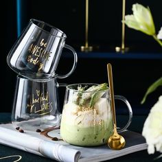 two mugs filled with green liquid sitting on top of a book next to flowers