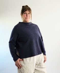 a woman standing in front of a white wall wearing a blue sweater and beige pants