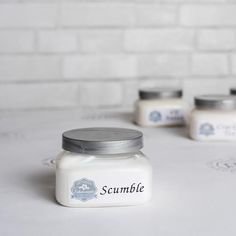 three small jars with labels on them sitting on a white tablecloth next to a brick wall