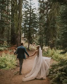 a bride and groom walking through the woods holding hands