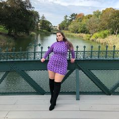 a woman in a purple dress is standing on a bridge near the water and trees