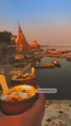 a person holding a bowl of food in front of a body of water with boats on it