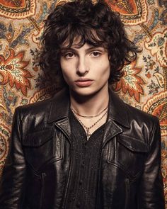 a man with long hair wearing a black leather jacket and standing in front of a floral wallpaper