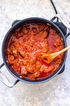 a pot filled with meat and sauce on top of a counter next to a wooden spoon