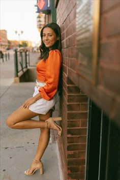 a woman leaning against a brick wall in an orange top and white skirt with her legs crossed