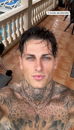 a man with tattoos on his chest sitting in front of a balcony looking at the camera