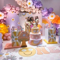 two women standing in front of a cake on a table with balloons and decorations around it