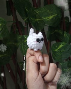 a hand holding a tiny crocheted white cat in front of a plant with snowflakes