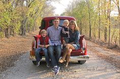 a family sitting in the back of an old pickup truck on a dirt road surrounded by trees