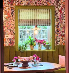 a dining room table with flowers on the wall and potted plants in the window