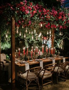 an outdoor dining table with candles and flowers hanging from it's ceiling, surrounded by greenery