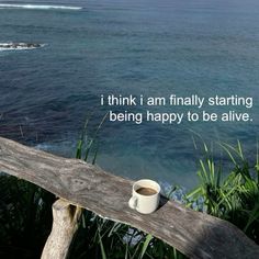 a cup of coffee sitting on top of a wooden bench next to the ocean with an inspirational quote