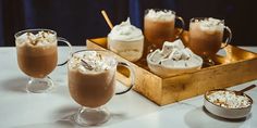 three glasses filled with hot chocolate and whipped cream on a table next to two bowls of marshmallows