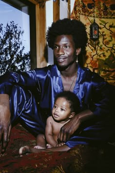 a man holding a baby in his lap while sitting on the floor next to a window