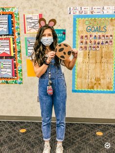 a woman wearing a face mask and overalls holds up a cookie in front of a bulletin board