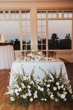 a table with white flowers and candles on it in front of large windows at dusk