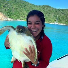 a woman holding a turtle in her arms on a boat with blue water and hills in the background