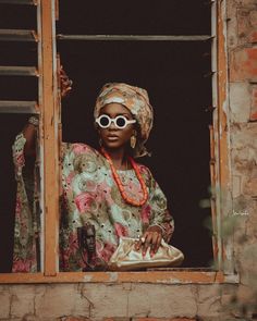 a woman wearing sunglasses and a head scarf looks out an open window at the camera