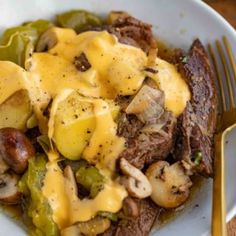 a white plate topped with meat and potatoes covered in cheese next to a fork on top of a table