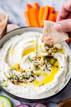 a hand holding a cracker over a bowl of hummus and carrots next to sliced cucumbers