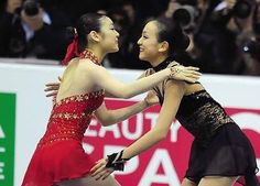 two women in red and black dresses dance together at an ice skating rink while people take pictures behind them