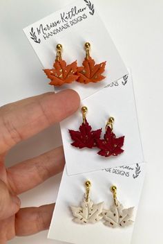 three pairs of maple leaf earrings are shown in different colors and sizes, one is gold, the other is red