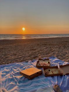 two pizzas are sitting on a blanket at the beach as the sun goes down