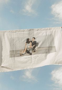 a man and woman sitting on top of a white banner flying in the blue sky