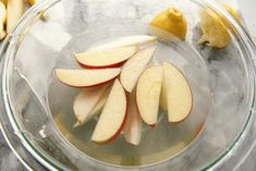 sliced apples in a glass bowl on a marble counter top next to bananas and lemon wedges