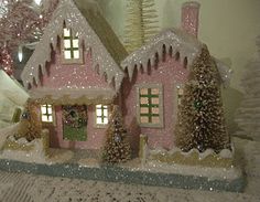 a small pink house with snow on the roof and trees in front of it, next to a christmas tree