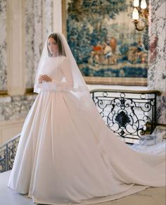 a woman in a white wedding gown and veil standing on a balcony with an ornate wallpapered background