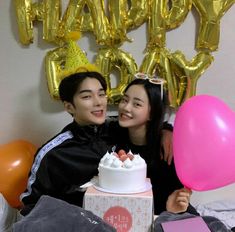 two people sitting next to each other in front of a cake and balloons with the words happy birthday written on it