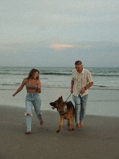 a man and woman walking their dog on the beach at sunset with one holding his leash