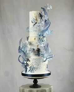 a three tiered cake with blue flowers on top sits on a pedestal in front of a gray background