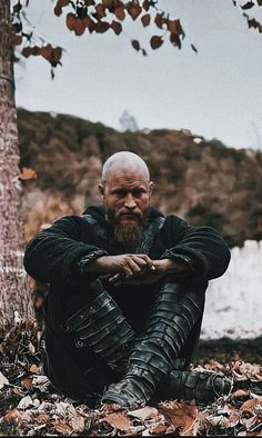 a man sitting on the ground in front of a tree with his hands folded up