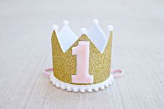 "* QUICK SHIP 3-5 days MINI CROWN: It is a mini crown in sparkly gold with white felt liner, decorated with a pink and white 1, trimmed with mini white pom poms along bottom and on crown points. Attached to a pink 1/8\" elastic- to be worn comfortably like a headband. Diameter of crown is 2.75\", and height 2.25\". *Note: Last picture is just to show example of size on a 1 year old. That crown can be found in my shop. CAKE TOPPER: Gold glitter \"one\" attached to a twine with pink/white poms on First Birthday Crown, Mini Crown, Birthday Gold, Girl 1st Birthday, Gold And Pink, Birthday Crown, My Son Birthday, Glitter Gold, Gold Birthday