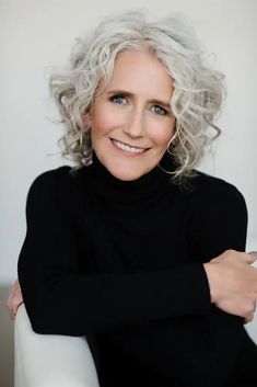 an older woman with white hair sitting in a chair and smiling at the camera while wearing a black turtle neck sweater